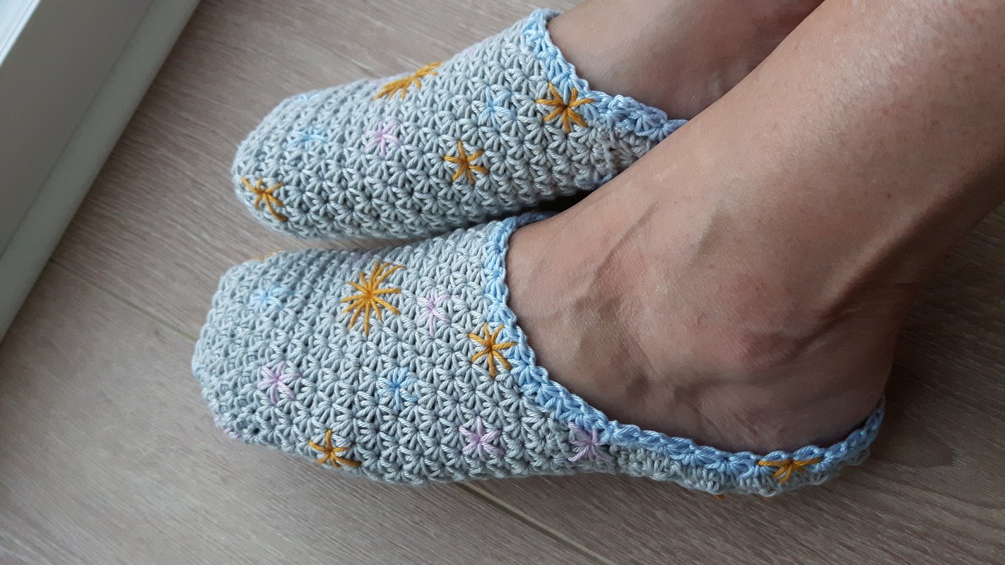 Nahaufnahme von Füßen in hellgrauen gehäkelten Hausschuhen mit gelben, hellblauen und rosa Sternenstickereien und hellblauem Rand auf hellem Holzboden.
