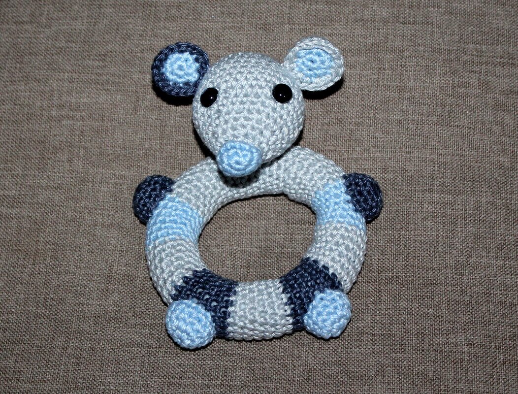 Gehäkelter Maus‑Greifring in Blau‑ und Grautönen, ringförmig mit angenähtem Mäusekopf und schwarzen Augen.