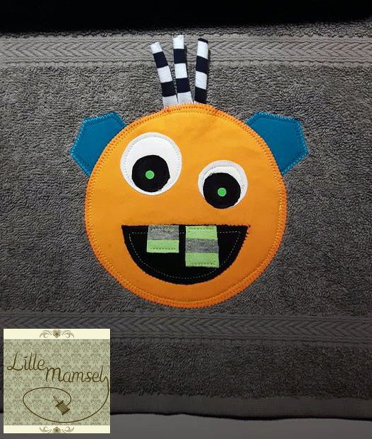Orange Monster-Applikation mit großen Augen, blauen Ohren und geringelten Fühlern auf grauem Handtuch