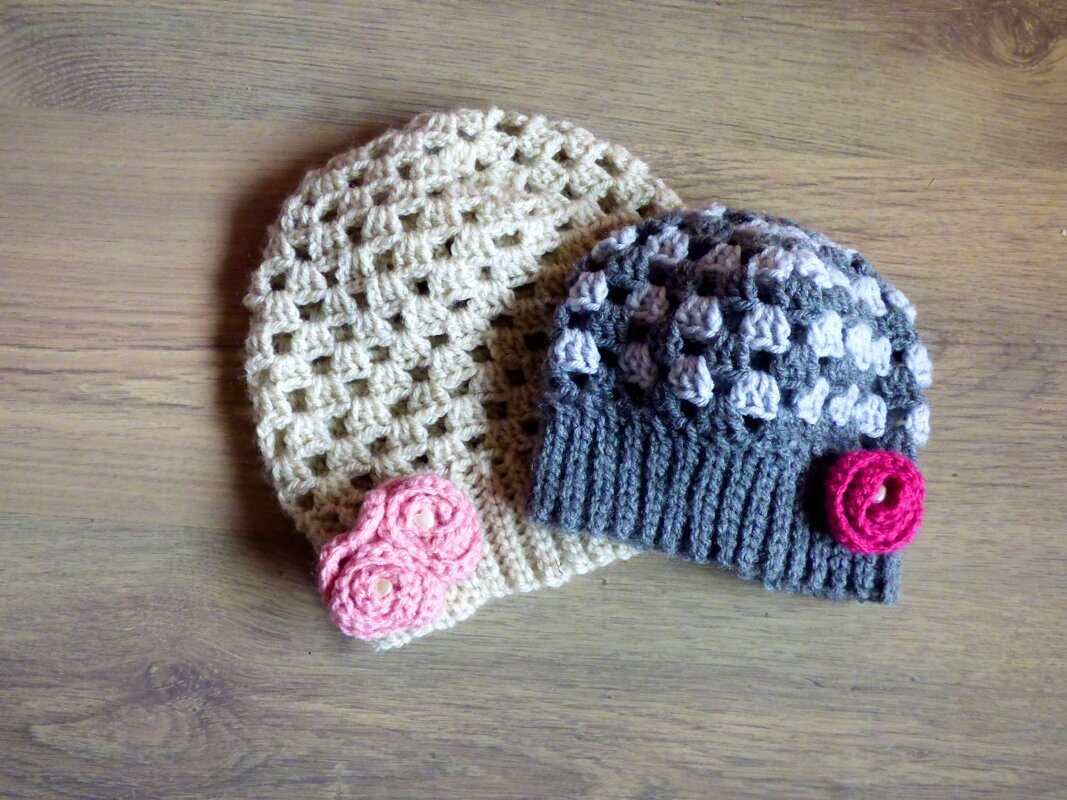 Zwei gehäkelte Mützen auf Holz: eine cremefarbene mit rosa Rosen, eine graue mit pinker Rose.