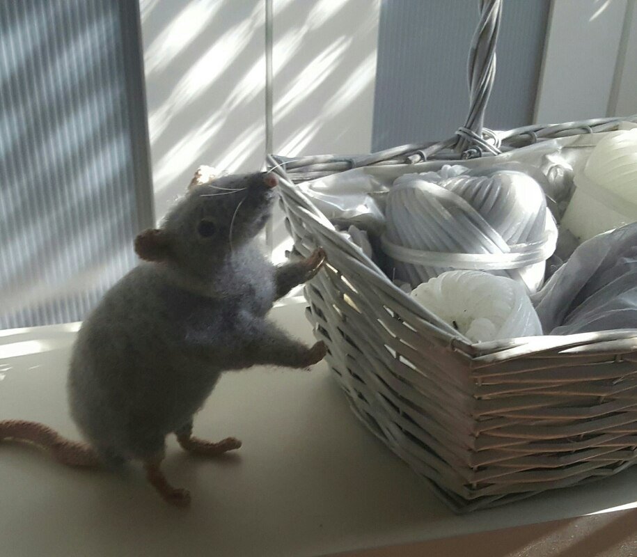 graue Amigurumi-Ratte lehnt an einem Korb mit Garnknäueln auf einem hellen Fensterbrett