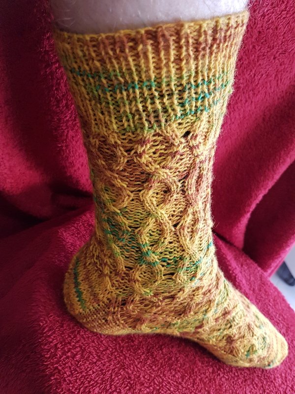 Handgestrickte gelbe Socke mit Zopfmuster und grünen Akzenten, getragen auf rotem Stoff