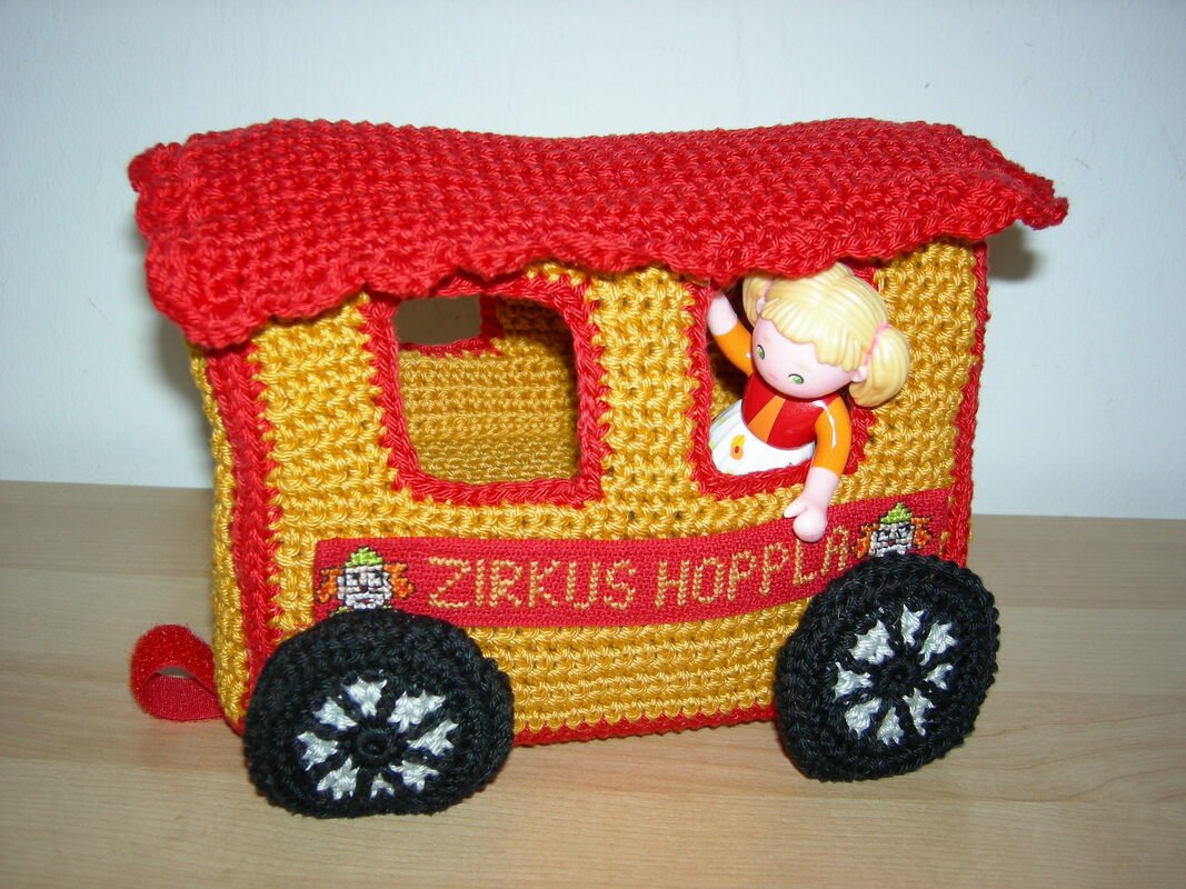 Gehäkelter gelb-roter Zirkuswagen mit schwarzen Rädern und einer kleinen Puppe im Fenster.