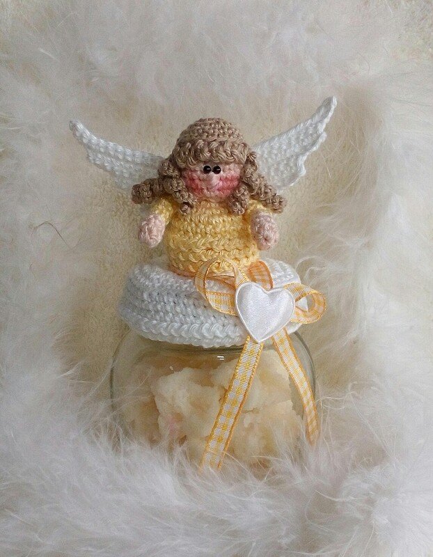 Gehäkelter Engel in gelbem Kleid mit weißen Flügeln auf dekoriertem Glas vor weißem Federhintergrund