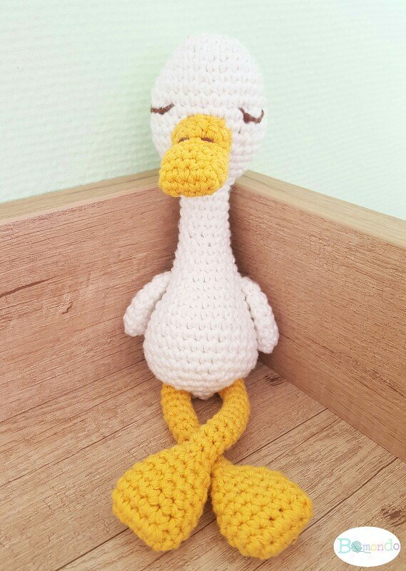 Gehäkelte weiße Ente mit gelbem Schnabel, verschränkten gelben Füßen und gestickten geschlossenen Augen, sitzt in einer Holzecke
