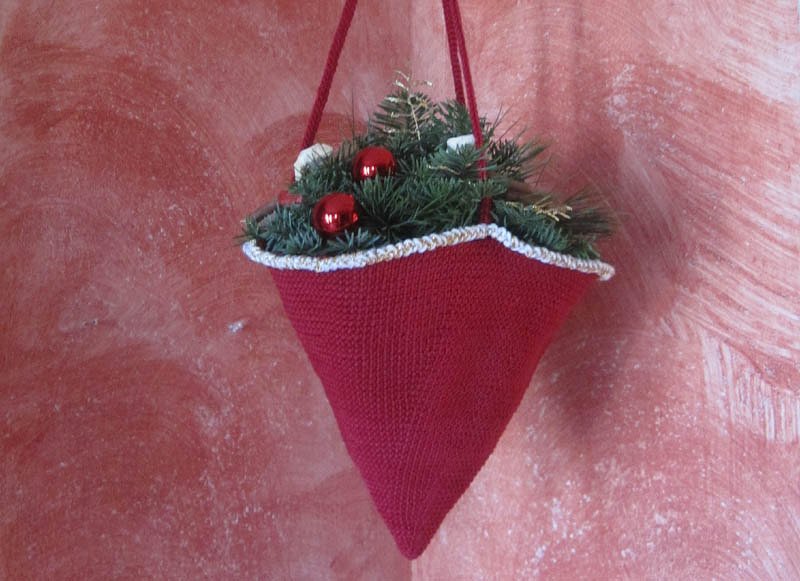 Konischer roter gehäkelter Hängebeutel mit weißem Rand, gefüllt mit Tannenzweigen und roten Weihnachtskugeln, an einer Aufhängung hängend