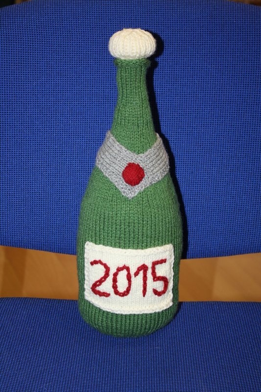 Gestrickte grüne Sektflasche mit weißem Flaschenkorken, grauem Kragen und Etikett '2015'.