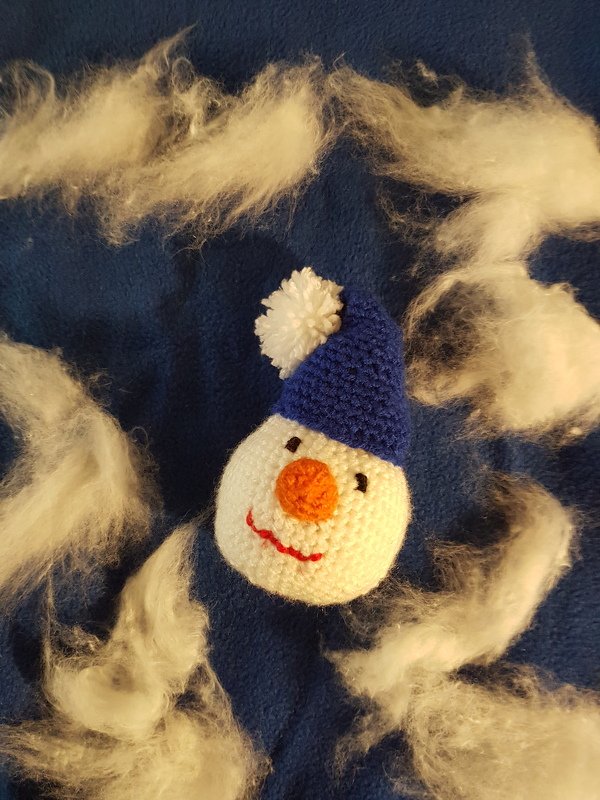 Gehäkelter weißer Schneemannkopf mit orangefarbener Nase und blauer Bommelmütze auf dunkelblauem Stoff, umgeben von weißen Fasern