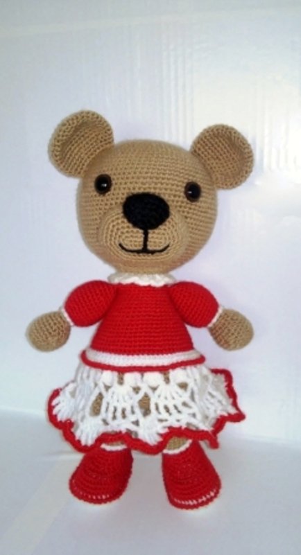 Gehäkelter beiger Amigurumi-Teddybär mit rotem Oberteil, weißem Spitzenrock und roten Schuhen, vor einem hellen, neutralen Hintergrund.