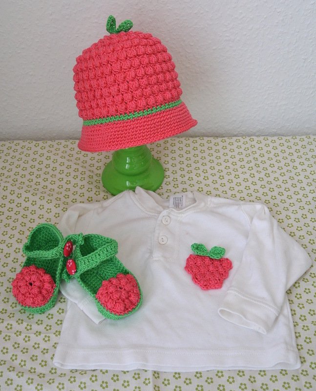 Gehäkeltes Baby-Set mit rot-grüner Mütze, grünen Riemchenschühchen und Erdbeer-Applikation auf weißem Shirt