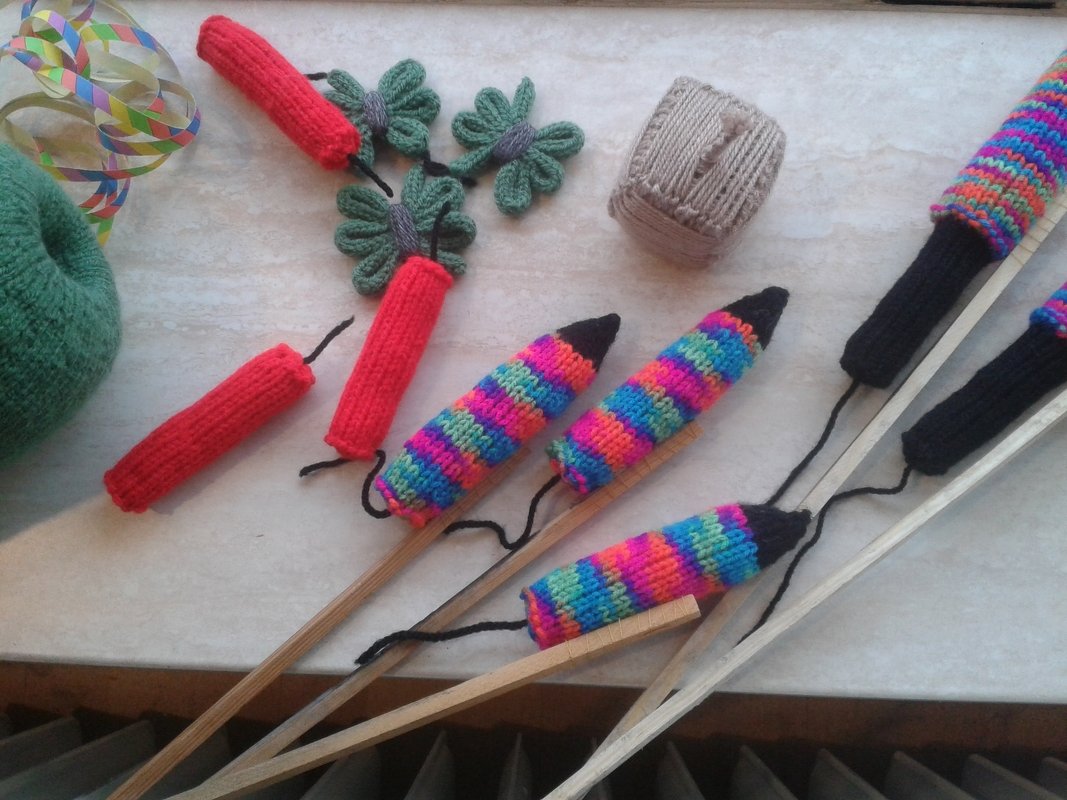 Mehrere handgestrickte bunte Hüllen in Raketen- und Zylinderform auf Holzstäben, grünes Kleeblatt, Garnknäuel und Papierband auf hellem Untergrund