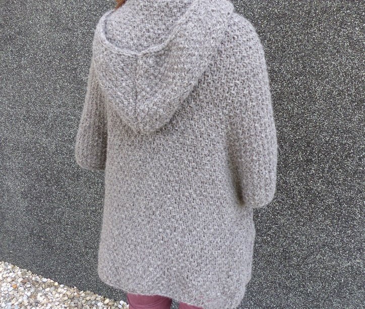 Rückansicht einer Person in einer hellgrauen, gestrickten Jacke mit Kapuze und dreiviertel Ärmeln vor einer dunklen Wand.