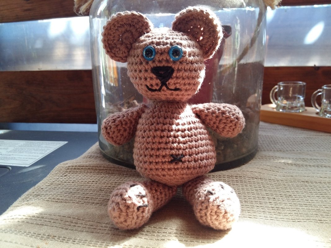 Hellbrauner gehäkelter Amigurumi-Teddybär mit blauen Knopfaugen auf beigem Tuch.