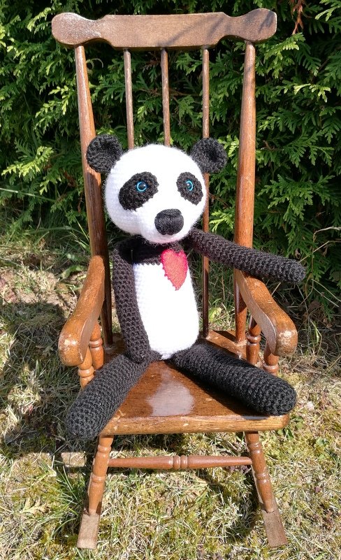 Gehäkelter Panda-Amigurumi mit blauen Augen und rotem Herz auf braunem Holzstuhl vor grünen Hecken