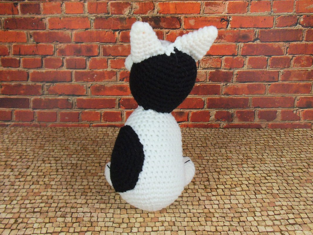 Schwarz-weißes Amigurumi-Kätzchen, Rückansicht vor roter Backsteinwand
