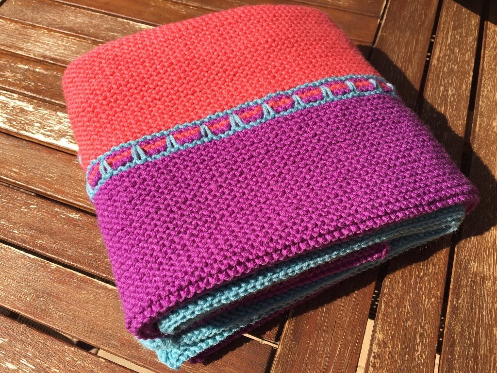 Gefaltete gestrickte Babydecke in Koralle, Lila und Türkis auf Holztisch.