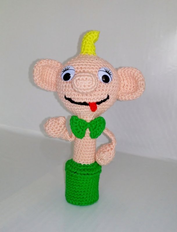 Gehäkelte Amigurumi-Figur mit Wackelaugen, gelber Haarlocke und grüner Fliege vor weißem Hintergrund