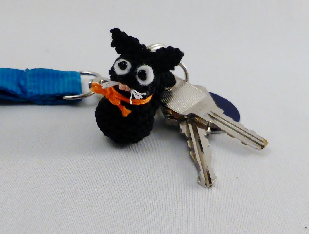 Gehäkelter schwarzer Katzen-Amigurumi als Schlüsselanhänger mit orangefarbener Schleife neben Schlüsseln