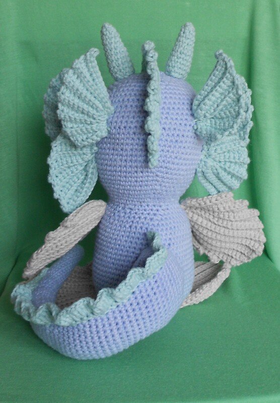 Rückansicht eines gehäkelten Amigurumi-Drachens in Hellblau mit mintgrünen Details und hellgrauen Flügeln vor grünem Hintergrund.