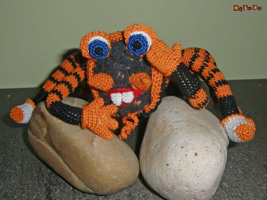 Gehäkelte orange‑schwarze Amigurumi‑Kreatur mit blauen Augen auf Steinen