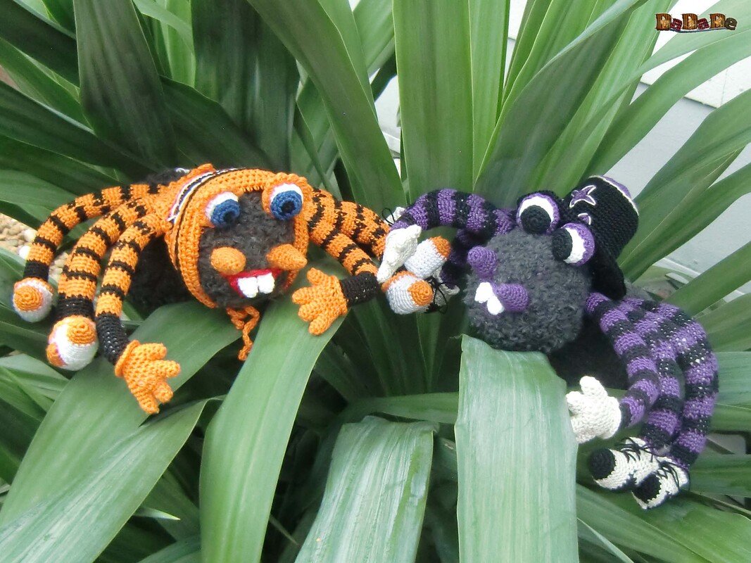 Zwei bunte Amigurumi-Spinnen in Orange-Schwarz und Lila-Schwarz auf großen grünen Blättern