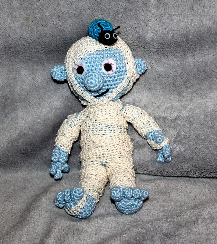 Hellblaue gehäkelte Mumienpuppe mit cremefarbenen Wicklungen und kleinem Käfer auf dem Kopf