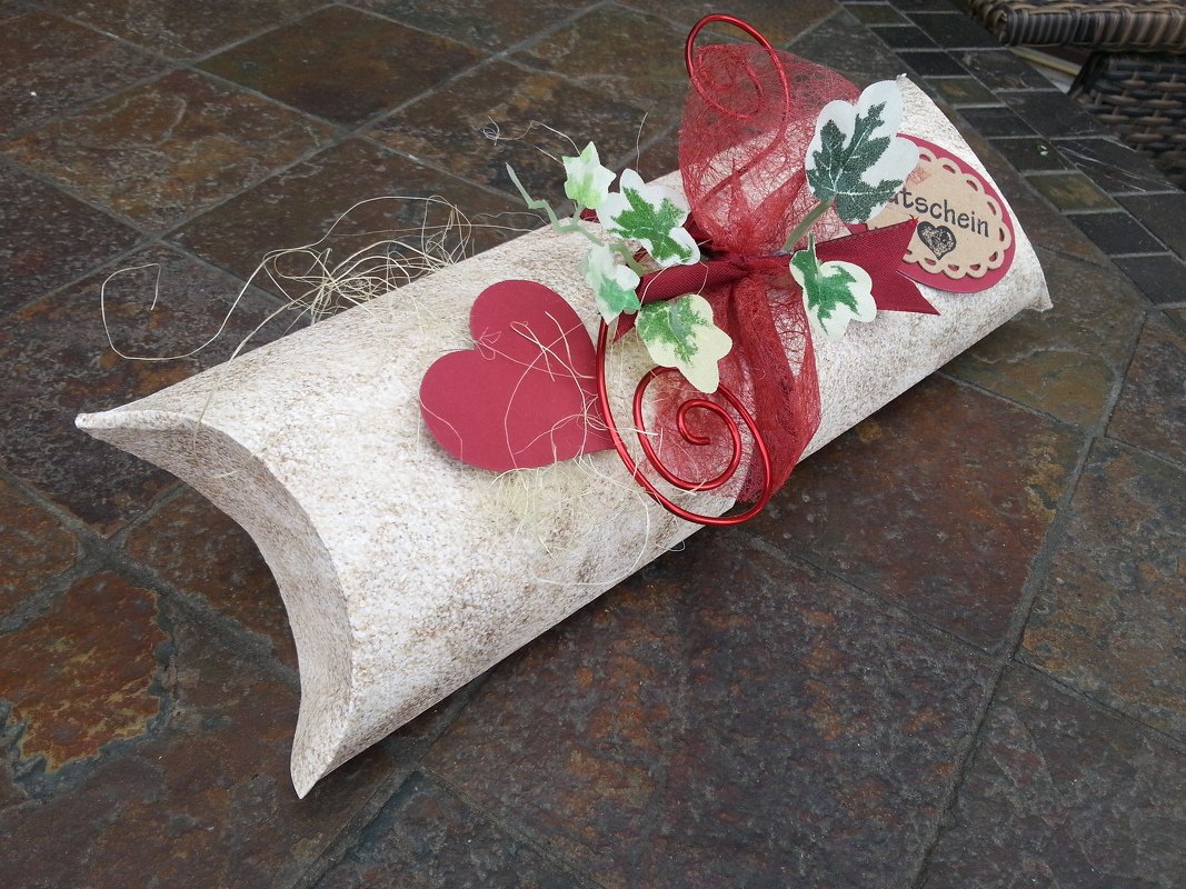DIY-Papier-Kissenbox mit rotem Herz, roter Schleife, Drahtspirale und Gutschein-Anhänger auf Steinboden