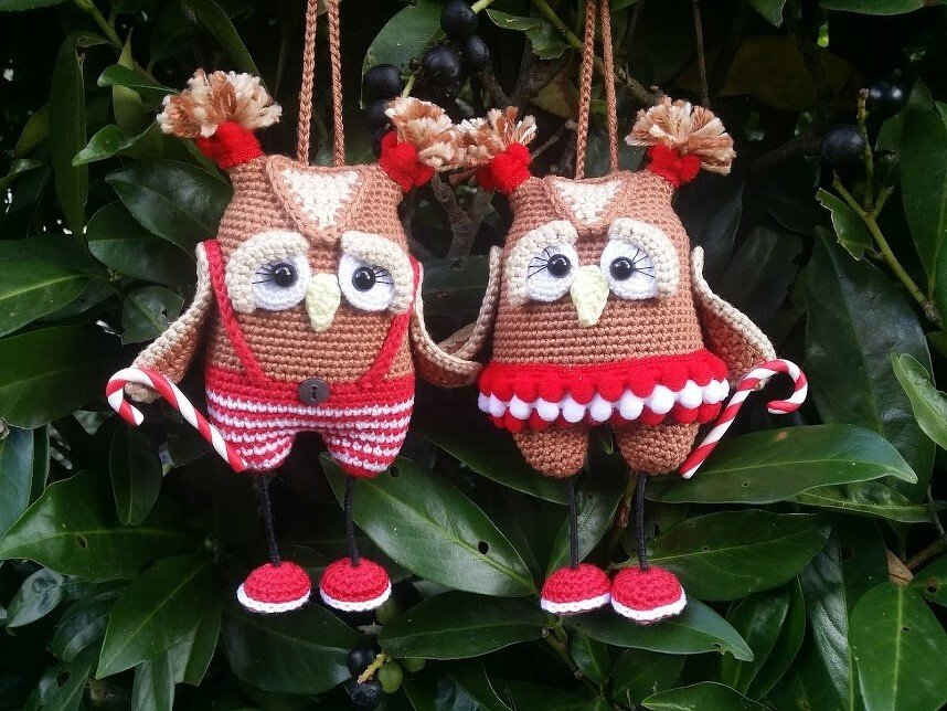 Zwei gehäkelte Amigurumi-Eulen mit rot-weißen Outfits und Zuckerstangen, hängend an grünen Blättern.