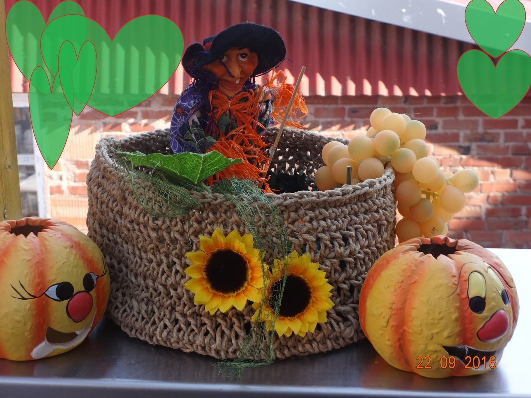 Gehäkelter Korb in Naturfarbe mit Sonnenblumen, einer Hexenpuppe, Trauben-Deko und Kürbisfiguren auf einem Tisch.