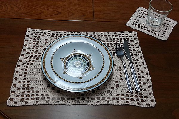 Häkel-Platzset im Filetmuster mit kleinem Untersetzer, Teller, Besteck und Glas auf Holztisch