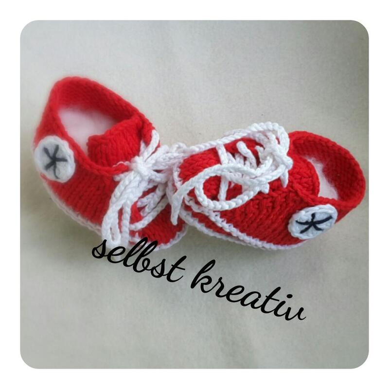Rote handgestrickte Babyschuhe im Sneaker‑Stil mit weißen Schnürsenkeln auf hellem Untergrund