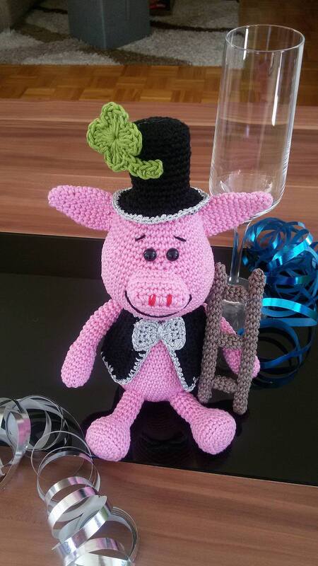 Rosa Amigurumi-Schwein mit Zylinder und Kleeblatt, trägt Weste und Fliege, neben einer kleinen Leiter und einem Sektglas