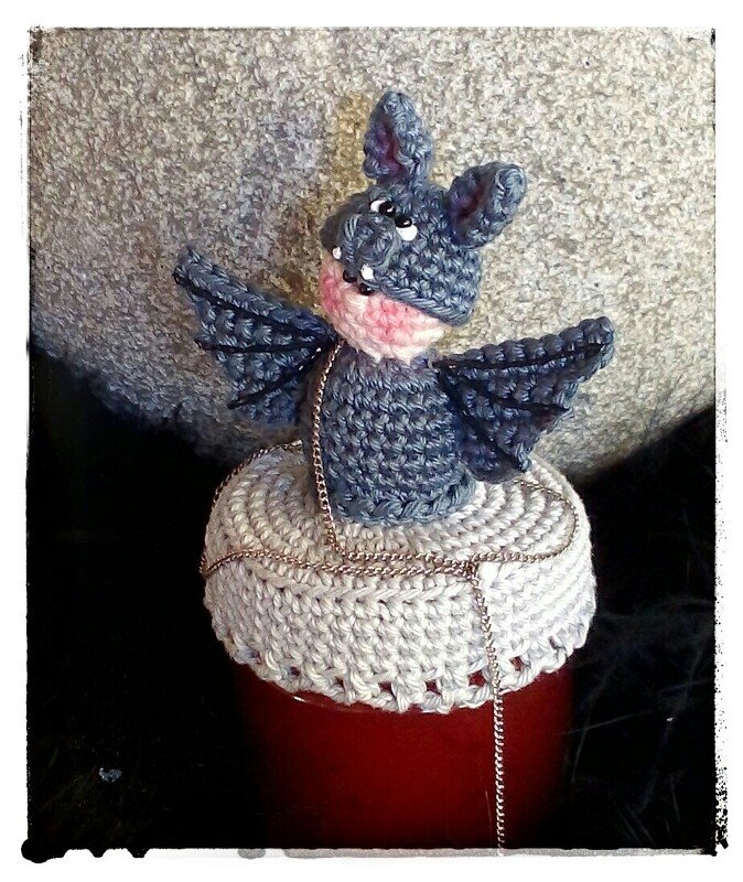 Gehäkelte graue Fledermaus-Amigurumi mit ausgebreiteten Flügeln auf weißem gehäkeltem Glasdeckel