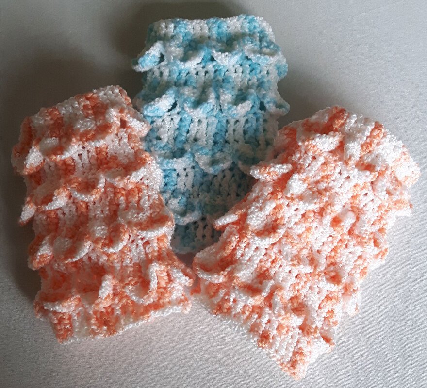 Drei gehäkelte Stulpen in Pastellfarben (Orange/Weiß, Hellblau/Weiß) mit schuppenartigem Muster auf hellem Untergrund