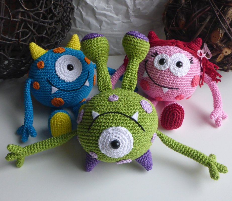Drei gehäkelte Amigurumi-Monster in grün, blau und rosa auf hellem Untergrund