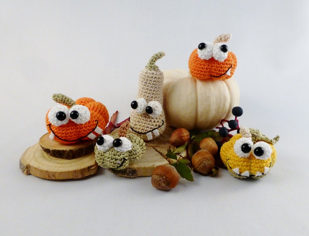 Gehäkelte Amigurumi-Kürbismonster mit großen Augen und Zähnen auf Holzscheiben, umgeben von Nüssen und einem weißen Kürbis.