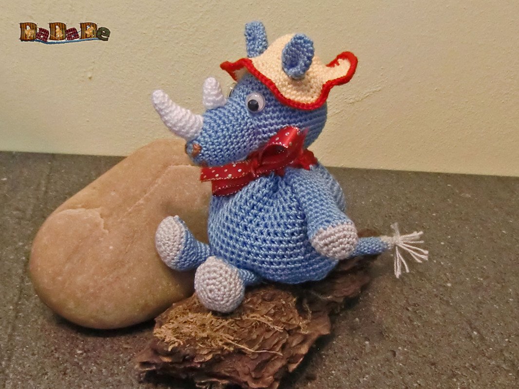 Blaues gehäkeltes Nashorn-Amigurumi mit weißem Horn, cremefarbenem Hut und roter Schleife, sitzend auf Naturmaterialien