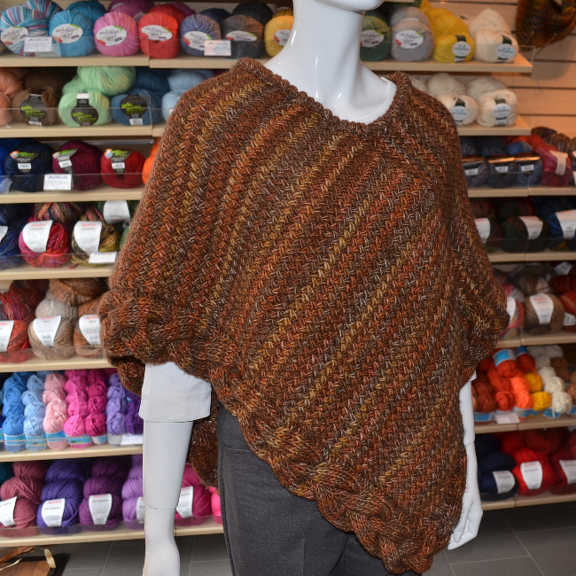 Braun-melierter Strickponcho mit gezopftem Saum auf Schaufensterpuppe vor Wollregal