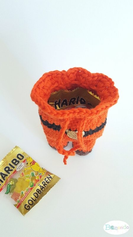 Nahaufnahme eines handgehäkelten orange-schwarzen Stiefels mit Kordelzug, gefüllt mit Bonbons; daneben eine kleine Tüte Gummibärchen.