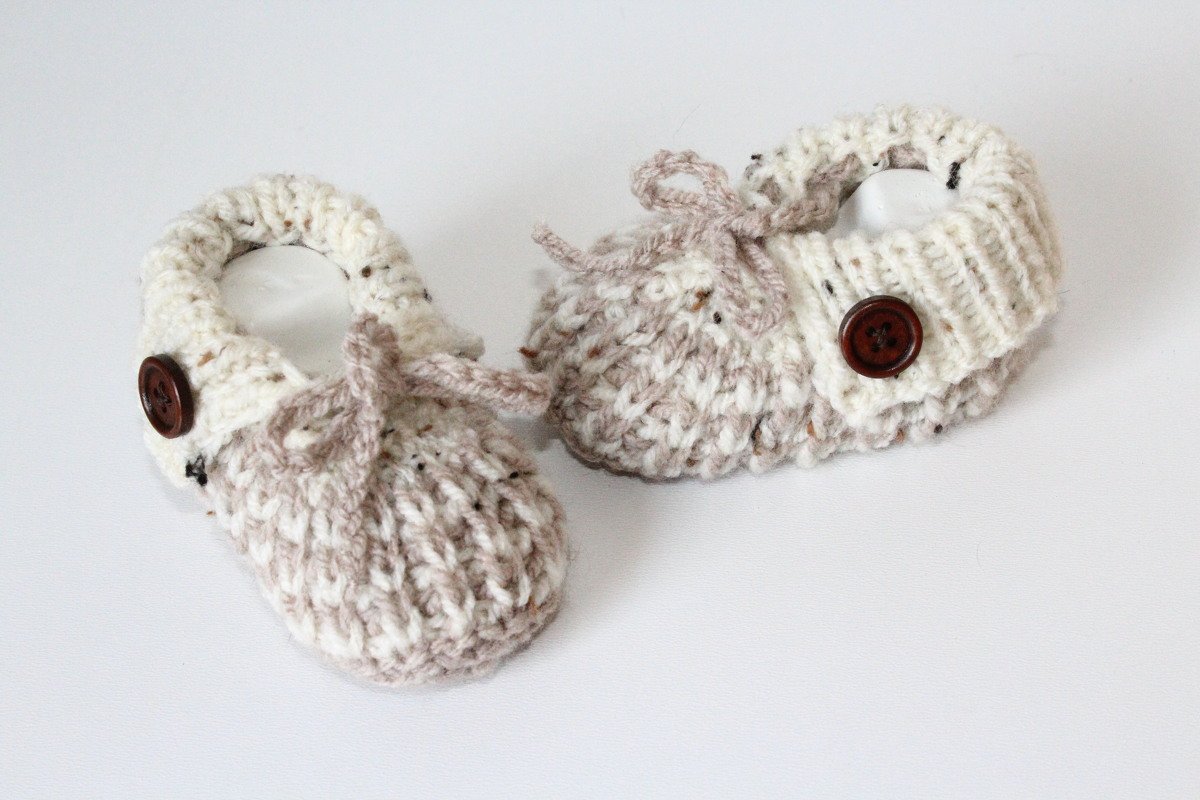 Gestrickte Babyschuhe in Creme und Beige mit braunen Knöpfen und Bindebändern auf hellem Hintergrund