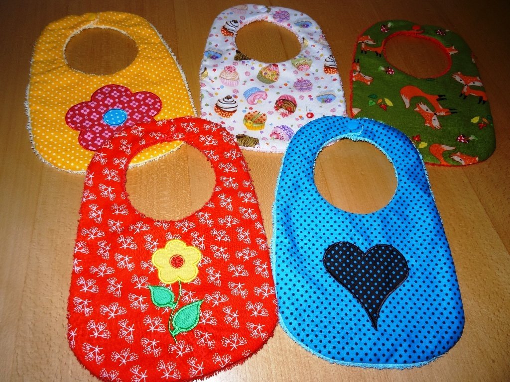 Fünf bunte Baby-Lätzchen mit unterschiedlichen Stoffmustern und applizierten Motiven auf einem Holztisch.