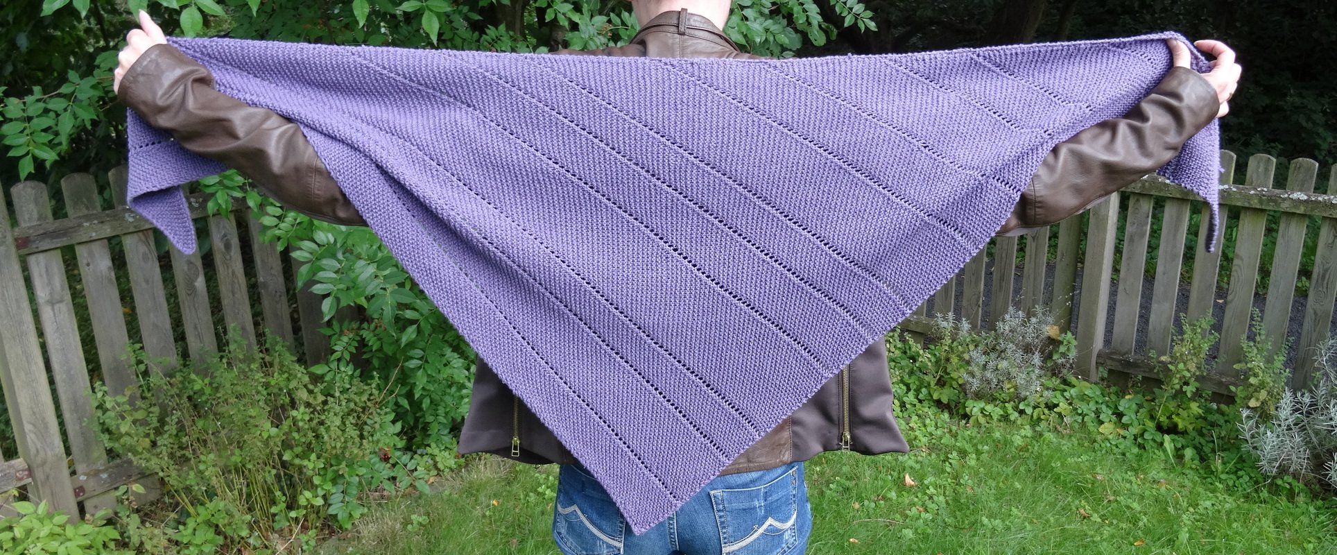 Großes gestricktes Dreieckstuch in Lila mit kraus-rechts-Muster und Reihen aus Löchern, gehalten von hinten