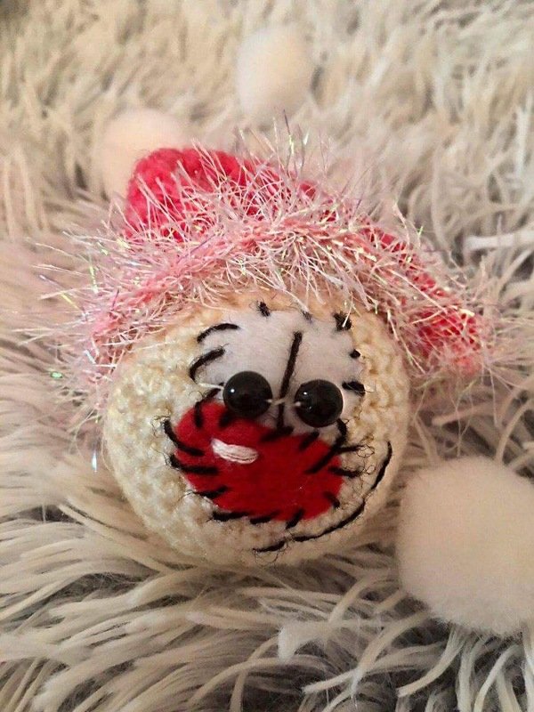 Runder gehäkelter Amigurumi-Schlüsselanhänger in Beige mit schwarzen Knopfaugen, rotem Mund und pinkem Glitzergarn auf weißem, flauschigem Stoff