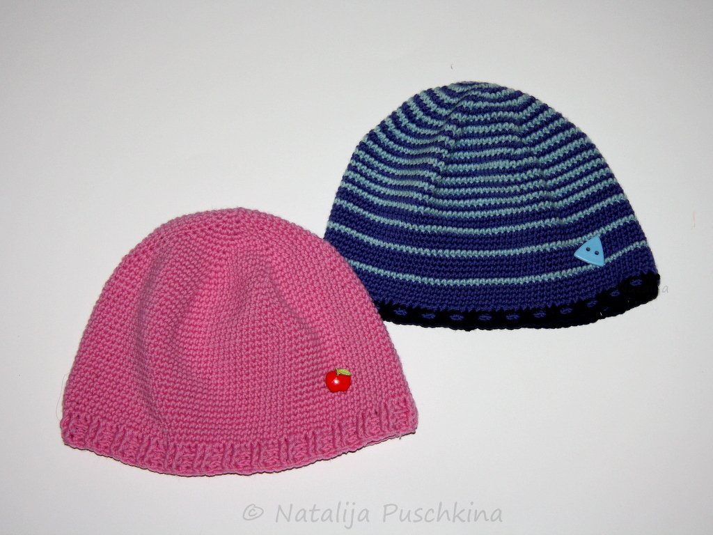 Zwei handgehäkelte Mützen: rosa Mütze mit Apfel-Applikation und blau-gestreifte Mütze mit dreieckigem Knopf.