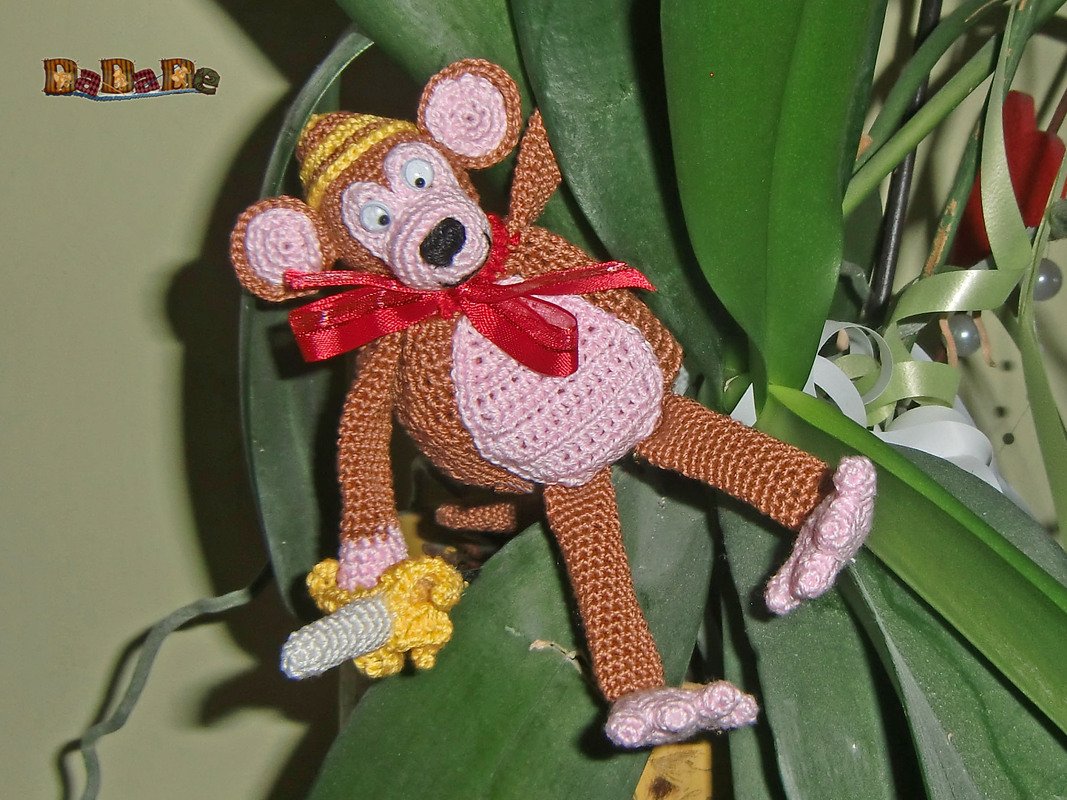 Gehäkelter brauner Amigurumi-Affe mit rosa Bauch, gelber Mütze, roter Schleife und kleiner Banane zwischen grünen Blättern