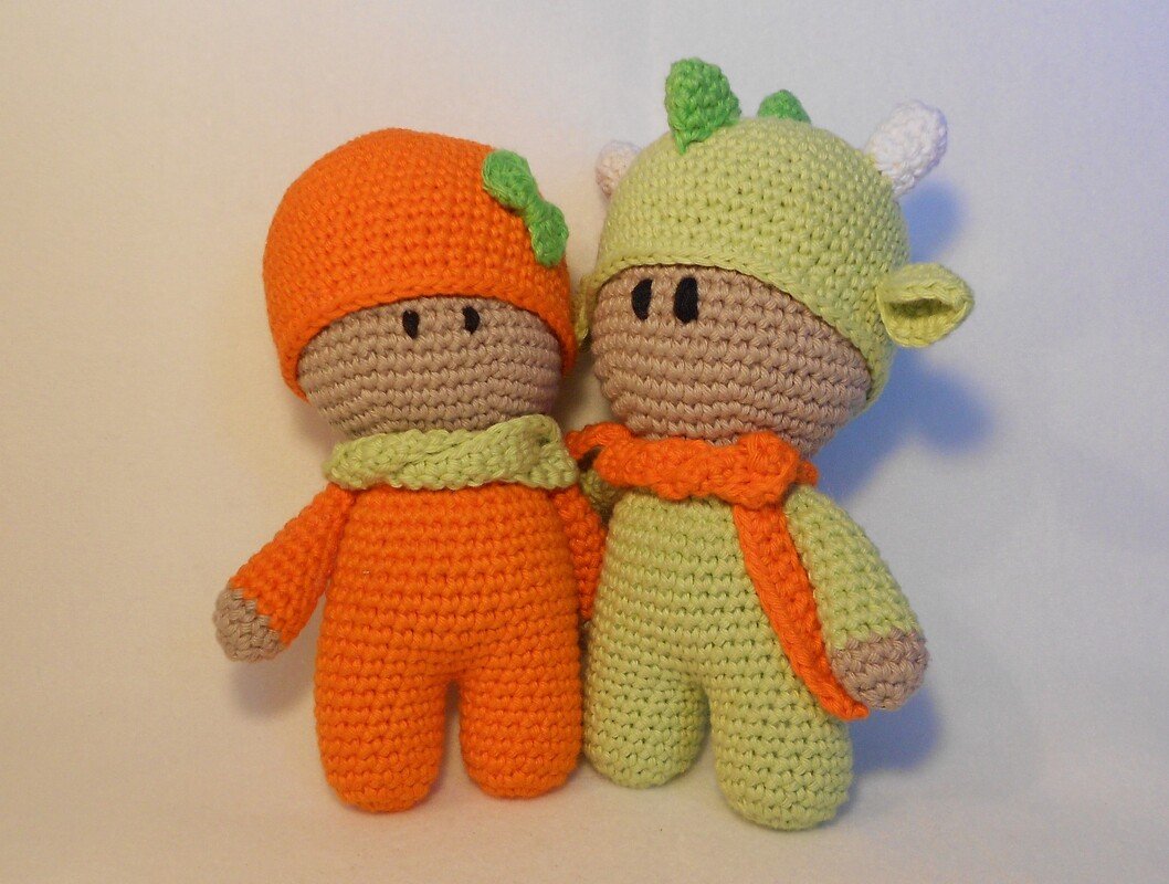 Zwei gehäkelte Amigurumi-Puppen nebeneinander, links orange mit Blattdetail an der Mütze, rechts hellgrün mit Hörnchen und Öhrchen, beide mit Schals