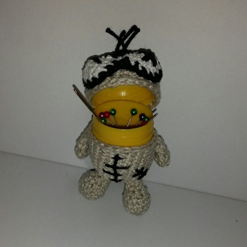 Beige gehäkeltes Skelett‑Amigurumi mit schwarzer Stickerei und geöffneter gelber Dose voller Stecknadeln