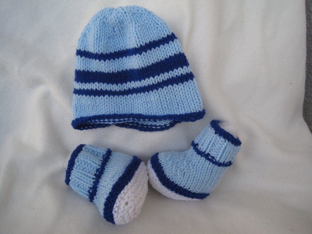 Gestrickte hellblaue Baby-Mütze mit dunklen Streifen und zwei passende Söckchen mit weißen Sohlen auf hellem Untergrund