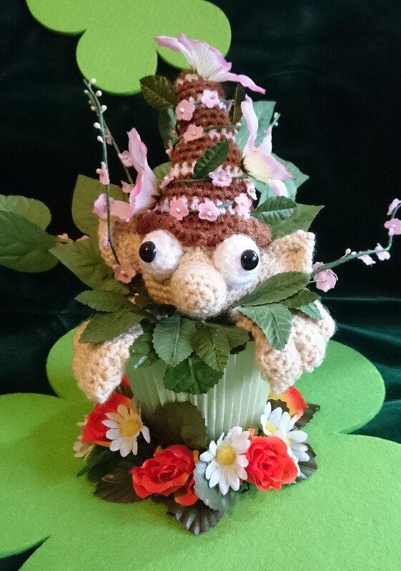 Gehäkeltes Amigurumi mit großen Augen im Blumentopf, umgeben von grünen Blättern und rosa künstlichen Blumen