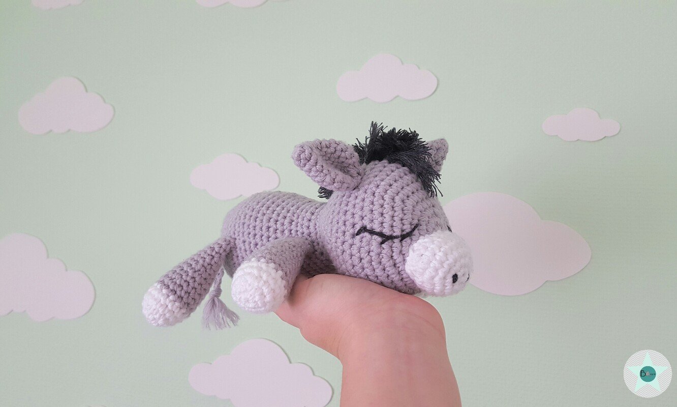 hellgrauer gehäkelter Amigurumi-Esel mit dunkler Mähne und weißen Akzenten, schlafend in einer Hand vor hellgrünem Hintergrund mit Wolken